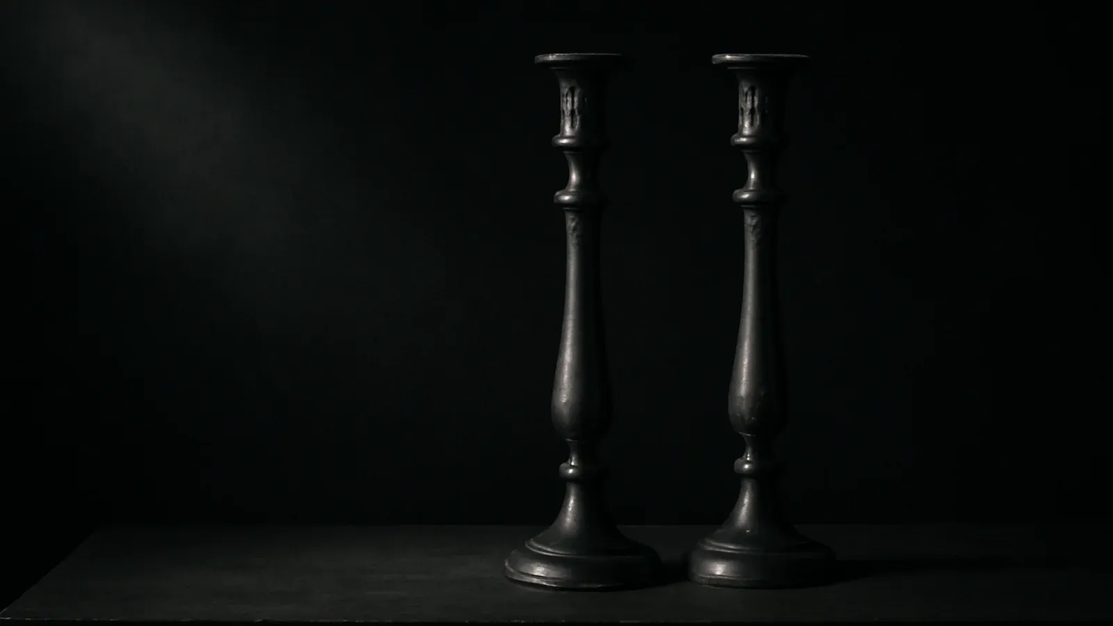 Two antique silver candlesticks standing in darkness, caught by a single shaft of light.